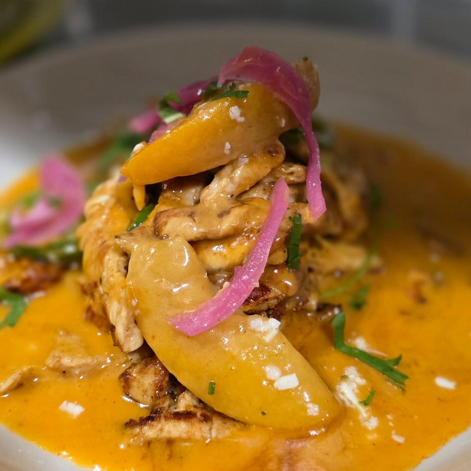 Close-up of seared meat and vegetables in orange sauce, garnished with pickled onions, herbs, and white crumbles.