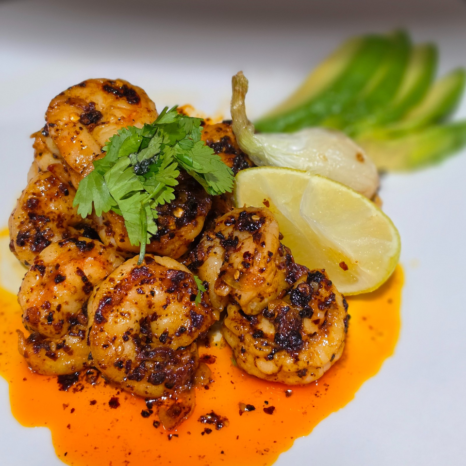 Grilled spiced shrimp garnished with cilantro, lemon wedges, and green peppers on a plate of orange chili oil.
