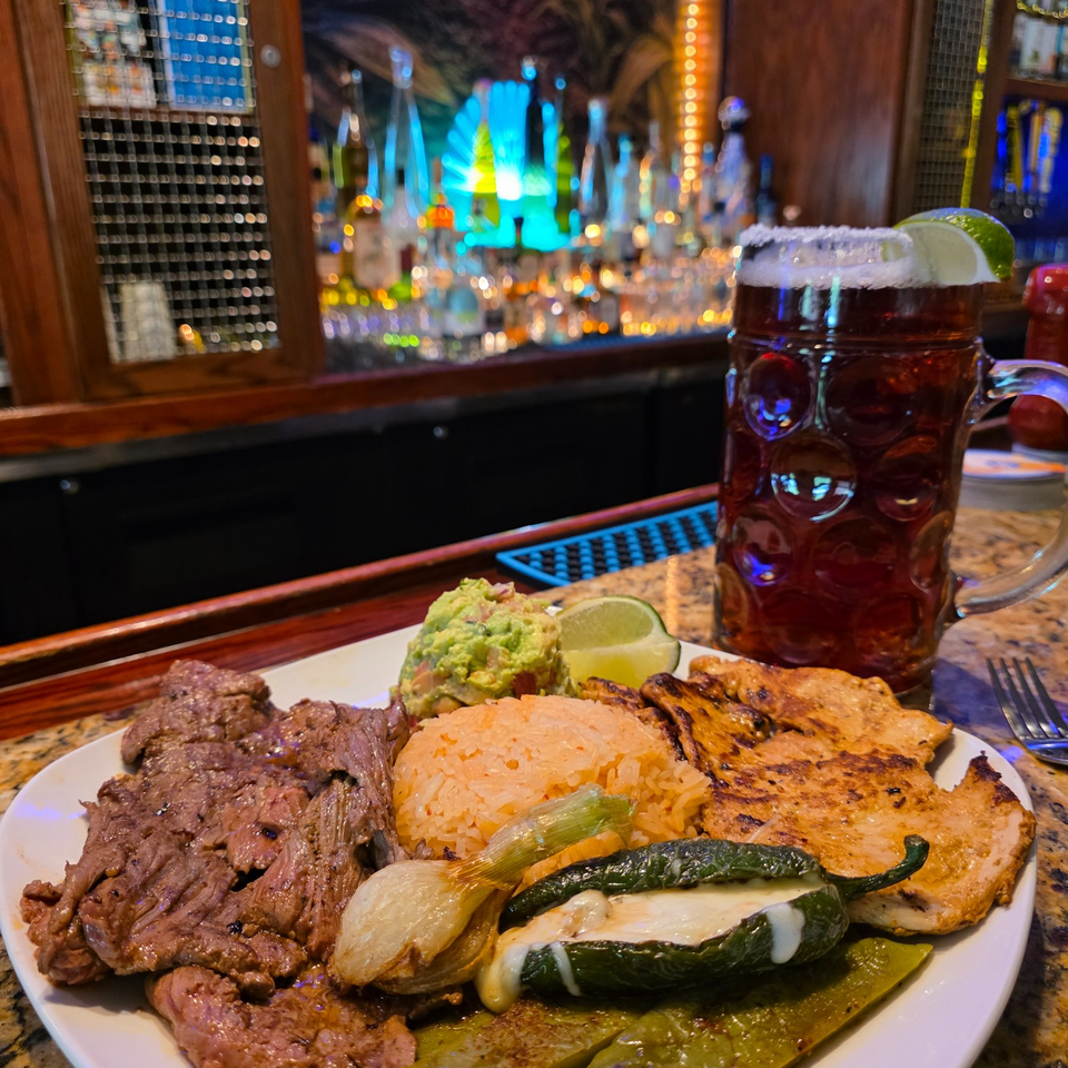 Grilled steak, chicken, rice, guacamole, roasted vegetables, and lime served with a foamy beer at a lively bar.